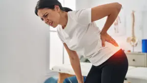 Mujer joven en un consultorio de fisioterapia, con expresión de dolor agudo, sosteniéndose la espalda baja. Un destello rojo resalta la zona afectada, indicando dolor ciático y planteando la pregunta implícita sobre qué hacer ciática.